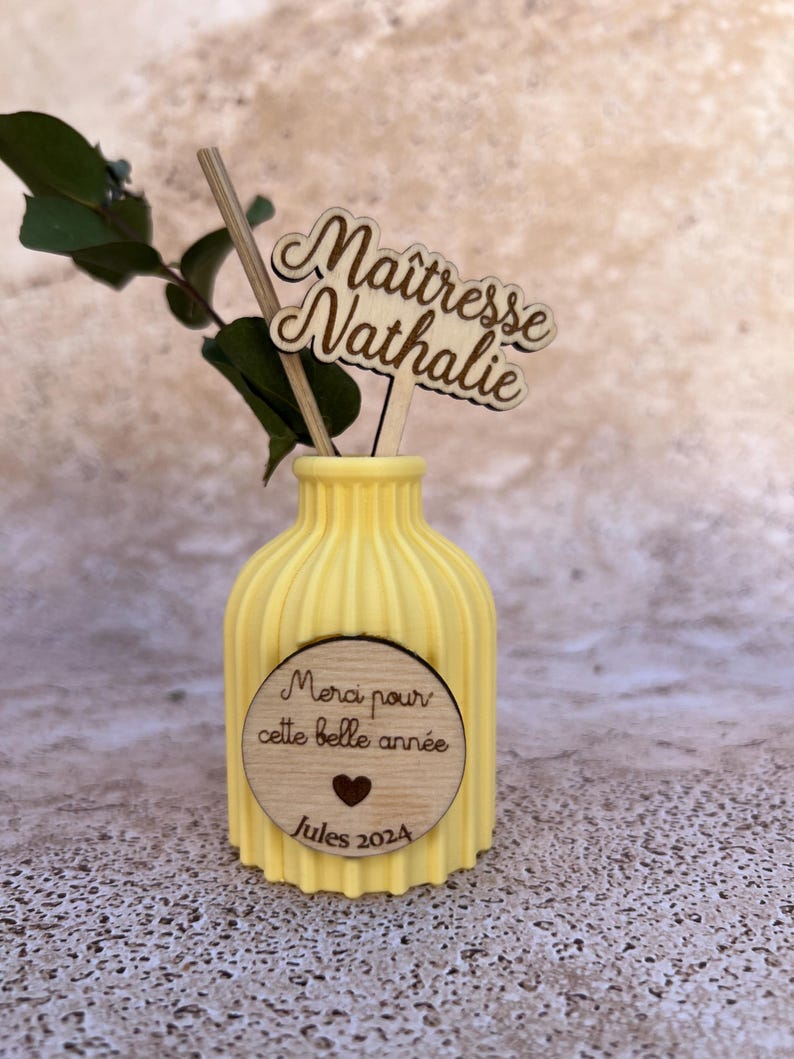 May include: A yellow ribbed vase featuring a circular wooden plaque with the text "Merci pour cette belle ann&eacute;e", a heart icon, and "Jules 2024". A wooden topper displays the text "Ma&icirc;tresse Nathalie" alongside a green eucalyptus sprig.