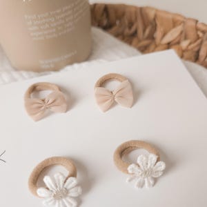 K&ouml;nnte beinhalten: Vier beige Baby-Haarspangen mit verschiedenen Designs liegen auf einer wei&szlig;en Oberfl&auml;che. Zwei Spangen haben ein Schleifendesign, zwei eine wei&szlig;e G&auml;nsebl&uuml;mchenblume. Der Text "tiful Bows MADE WITH LOVE" ist sichtbar.
