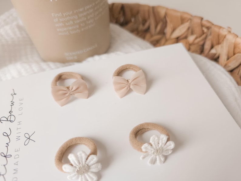 K&ouml;nnte beinhalten: Vier beige Baby-Haarspangen mit verschiedenen Designs liegen auf einer wei&szlig;en Oberfl&auml;che. Zwei Spangen haben ein Schleifendesign, zwei eine wei&szlig;e G&auml;nsebl&uuml;mchenblume. Der Text "tiful Bows MADE WITH LOVE" ist sichtbar.