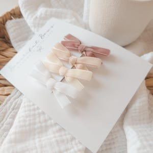 May include: A set of four velvet hair bows in white, cream, and dusty rose, arranged on a white card. The card is placed on a white, textured fabric, with a woven basket and a white ceramic cup in the background. The card reads "Beautiful Bows, Handmade with Love."