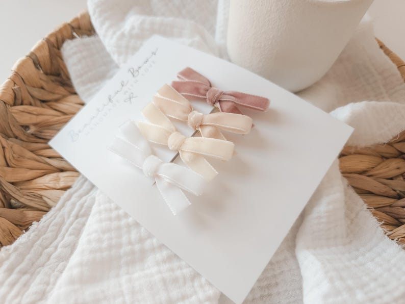 May include: A set of four velvet hair bows in white, cream, and dusty rose, arranged on a white card. The card is placed on a white, textured fabric, with a woven basket and a white ceramic cup in the background. The card reads "Beautiful Bows, Handmade with Love."