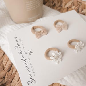 K&ouml;nnte beinhalten: Vier Baby-Haarb&auml;nder mit Schleifen und Blumenakzenten. Die Haarb&auml;nder liegen auf einer wei&szlig;en Karte mit dem Text "Beautiful Bows Handmade With Love". Die Schleifen sind hellbeige und die Blumen wei&szlig;.