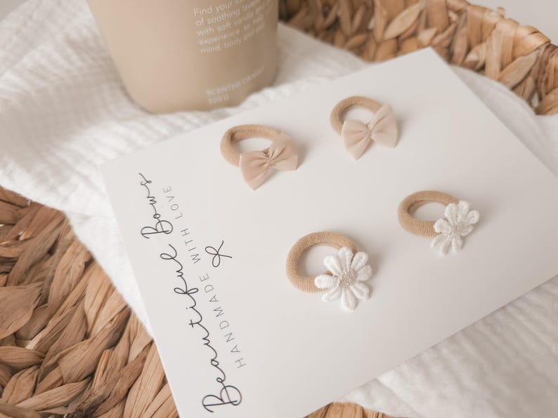 K&ouml;nnte beinhalten: Vier Baby-Haarb&auml;nder mit Schleifen und Blumenakzenten. Die Haarb&auml;nder liegen auf einer wei&szlig;en Karte mit dem Text "Beautiful Bows Handmade With Love". Die Schleifen sind hellbeige und die Blumen wei&szlig;.