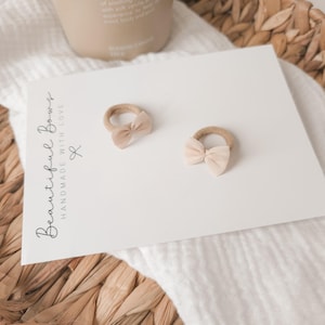 K&ouml;nnte beinhalten: Zwei beige Baby-Haarschleifen mit einer hellrosa Schleife auf einer wei&szlig;en Karte. Auf der Karte steht der Text "Beautiful Bows Handmade with Love". Die Schleifen befinden sich auf einer wei&szlig;en Oberfl&auml;che, mit einem geflochtenen Korb im Hintergrund.