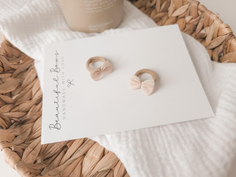 K&ouml;nnte beinhalten: Zwei beige Baby-Haarschleifen mit einer hellrosa Schleife auf einer wei&szlig;en Karte. Auf der Karte steht der Text "Beautiful Bows Handmade with Love". Die Schleifen befinden sich auf einer wei&szlig;en Oberfl&auml;che, mit einem geflochtenen Korb im Hintergrund.