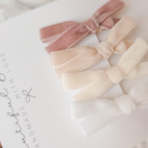 May include: Four velvet hair bows in shades of dusty rose, cream, and white are arranged on a white card. The bows are tied in a classic bow shape. The card has the text "Beautiful Bows, Handmade with Love."