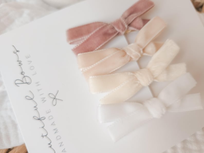 May include: Four velvet hair bows in shades of dusty rose, cream, and white are arranged on a white card. The bows are tied in a classic bow shape. The card has the text "Beautiful Bows, Handmade with Love."