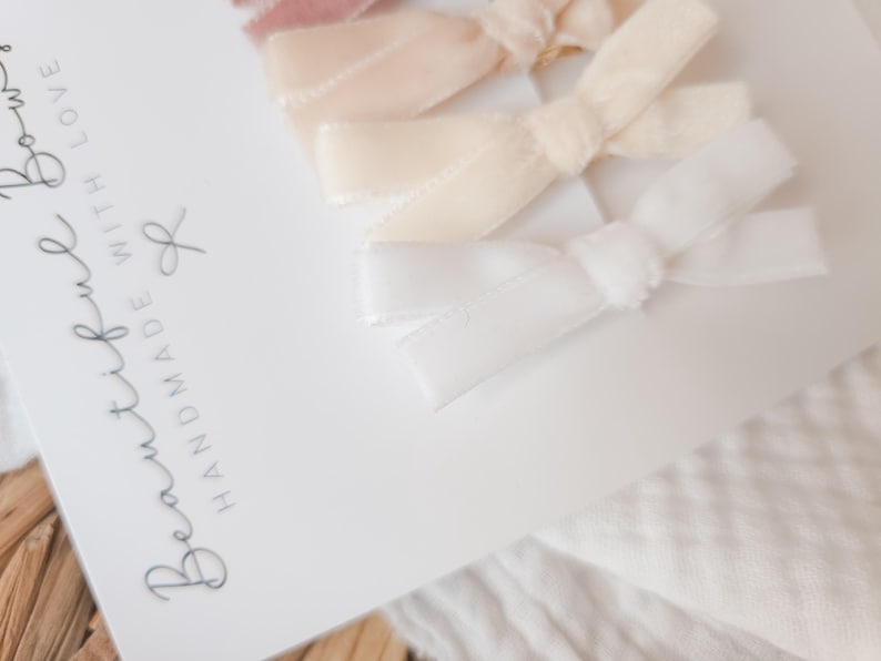 May include: A set of four velvet hair bows in shades of pink, cream, and white, arranged on a white card. The card has the text "Beautiful Bows Handmade With Love" in cursive. The bows are tied in a classic bow shape.