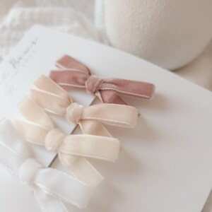 May include: A collection of velvet hair bows in shades of white, cream, and mauve. The bows are arranged on a white card, with a soft, neutral background. These accessories are ideal for adding a touch of elegance to any hairstyle.