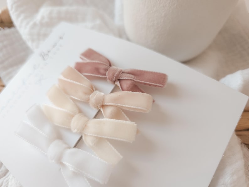 May include: A collection of velvet hair bows in shades of white, cream, and mauve. The bows are arranged on a white card, with a soft, neutral background. These accessories are ideal for adding a touch of elegance to any hairstyle.