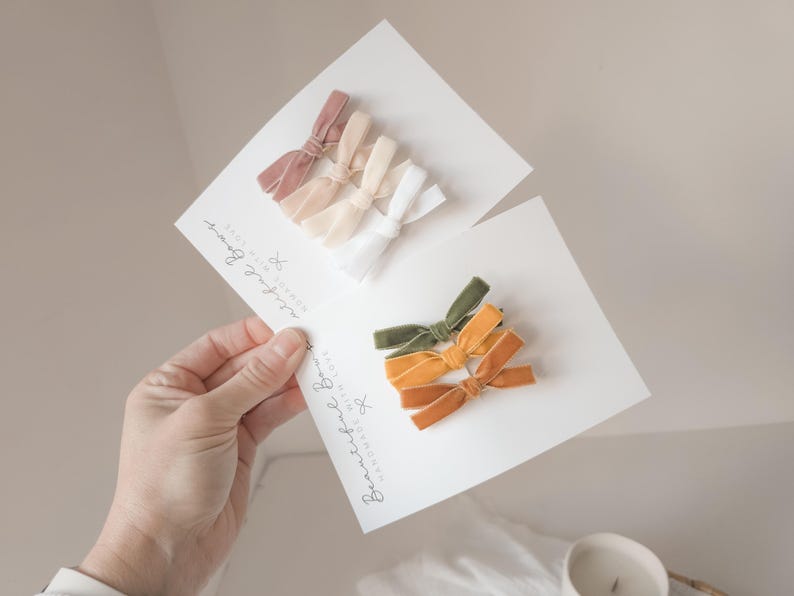 May include: Two sets of handmade velvet bows in various colors, including pink, cream, white, olive green, and orange. Each set is displayed on a white card with the text "Beautiful Bows" in cursive.