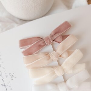 May include: A set of four velvet hair bows in varying shades of pink and cream. The bows are arranged on a white card with the words "Beautiful Bows Made With Love" in cursive script. The bows are tied in a classic bow shape.