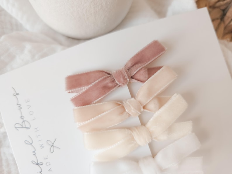 May include: A set of four velvet hair bows in varying shades of pink and cream. The bows are arranged on a white card with the words "Beautiful Bows Made With Love" in cursive script. The bows are tied in a classic bow shape.