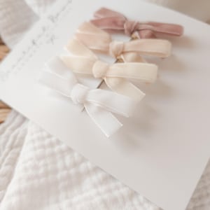 May include: Four velvet hair bows in shades of white, cream, and mauve are arranged on a white card. The bows are tied in a classic bow shape. The card is placed on a white, textured surface, with a woven basket in the background.
