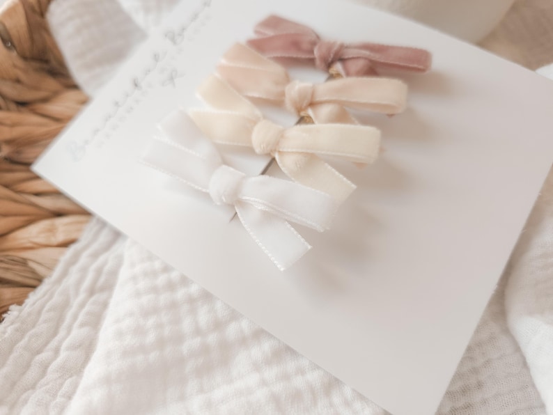 May include: Four velvet hair bows in shades of white, cream, and mauve are arranged on a white card. The bows are tied in a classic bow shape. The card is placed on a white, textured surface, with a woven basket in the background.