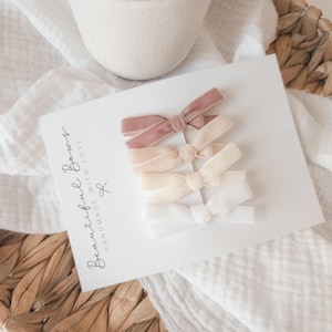 May include: A set of four velvet hair bows in varying shades of pink and white, arranged on a white card. The card has the text "Beautiful Bows Handmade With Love". The bows are displayed on a white fabric and a woven basket.