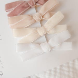 May include: Four velvet hair bows in shades of pink, cream, and white are arranged on a white card. The bows are tied in a classic bow shape. The card has the words "Beautiful Handmade With Love" printed on it.