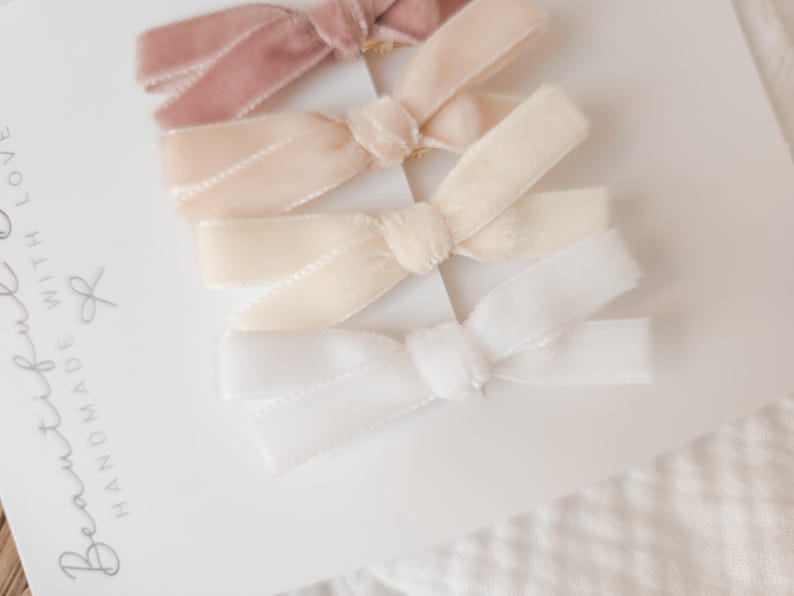 May include: Four velvet hair bows in shades of pink, cream, and white are arranged on a white card. The bows are tied in a classic bow shape. The card has the words "Beautiful Handmade With Love" printed on it.