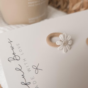K&ouml;nnte beinhalten: Zwei beige Haargummis mit wei&szlig;en G&auml;nsebl&uuml;mchen-Verzierungen werden auf einer wei&szlig;en Karte pr&auml;sentiert. Auf der Karte steht der Text "ful Bows MADE WITH LOVE". Im Hintergrund befindet sich eine beige Flasche.