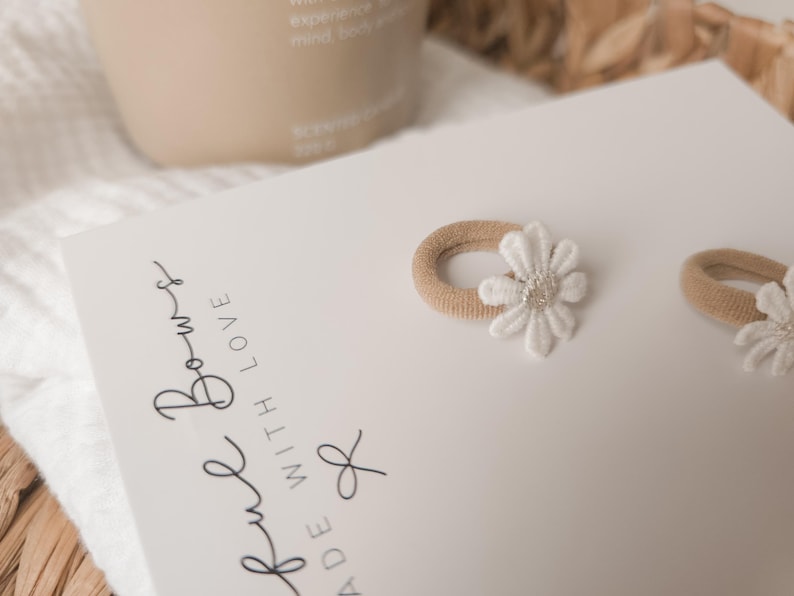 K&ouml;nnte beinhalten: Zwei beige Haargummis mit wei&szlig;en G&auml;nsebl&uuml;mchen-Verzierungen werden auf einer wei&szlig;en Karte pr&auml;sentiert. Auf der Karte steht der Text "ful Bows MADE WITH LOVE". Im Hintergrund befindet sich eine beige Flasche.