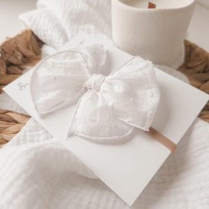 May include: White cotton bow with a dotted pattern, attached to a nude headband. The bow is displayed on a white surface with a wicker basket in the background.