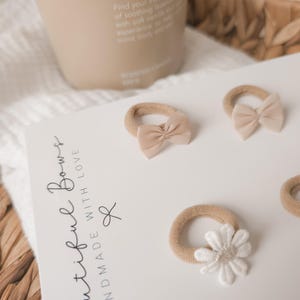 K&ouml;nnte beinhalten: Vier beige Haargummis mit Schleifen und einer wei&szlig;en Blume auf einer wei&szlig;en Oberfl&auml;che. Der Text "Beautiful Bows Handmade With Love" ist auf eine wei&szlig;e Karte gedruckt. Eine beige Kerze und ein Weidenkorb befinden sich im Hintergrund.