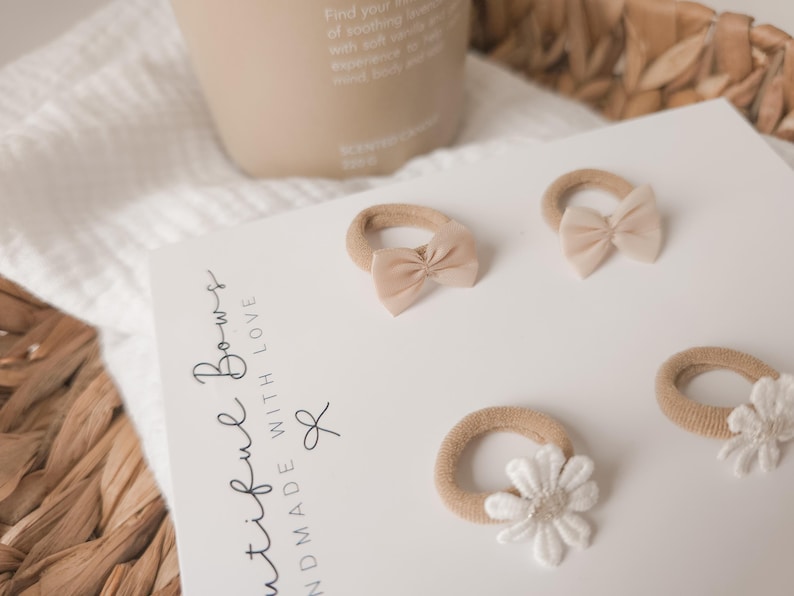 K&ouml;nnte beinhalten: Vier beige Haargummis mit Schleifen und einer wei&szlig;en Blume auf einer wei&szlig;en Oberfl&auml;che. Der Text "Beautiful Bows Handmade With Love" ist auf eine wei&szlig;e Karte gedruckt. Eine beige Kerze und ein Weidenkorb befinden sich im Hintergrund.