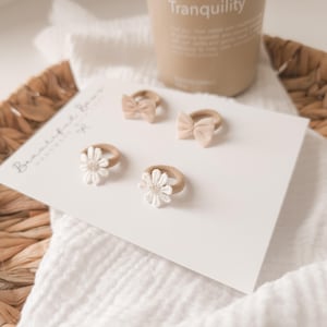 K&ouml;nnte beinhalten: Pr&auml;sentation von Baby-Haarschmuck, darunter zwei kleine Schleifen und zwei G&auml;nsebl&uuml;mchen-Designs, auf einer wei&szlig;en Karte. Die Karte befindet sich in einem Weidenkorb mit einem wei&szlig;en Tuch. Eine Kerze mit dem Wort "Tranquility" befindet sich im Hintergrund.