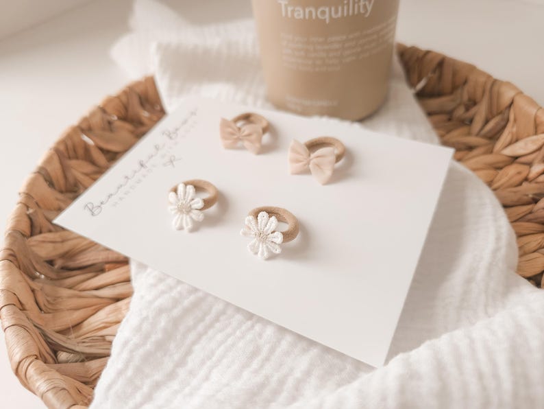 K&ouml;nnte beinhalten: Pr&auml;sentation von Baby-Haarschmuck, darunter zwei kleine Schleifen und zwei G&auml;nsebl&uuml;mchen-Designs, auf einer wei&szlig;en Karte. Die Karte befindet sich in einem Weidenkorb mit einem wei&szlig;en Tuch. Eine Kerze mit dem Wort "Tranquility" befindet sich im Hintergrund.