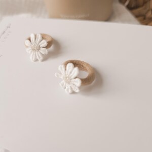 K&ouml;nnte beinhalten: Zwei Baby-Haargummis mit wei&szlig;en G&auml;nsebl&uuml;mchen-Akzenten. Die Haargummis haben ein beiges Gummiband und eine wei&szlig;e Blume mit einem Perlenmittelpunkt. Der Hintergrund ist eine wei&szlig;e Oberfl&auml;che mit den Worten "Soul Bows Made With Love" in Schreibschrift.