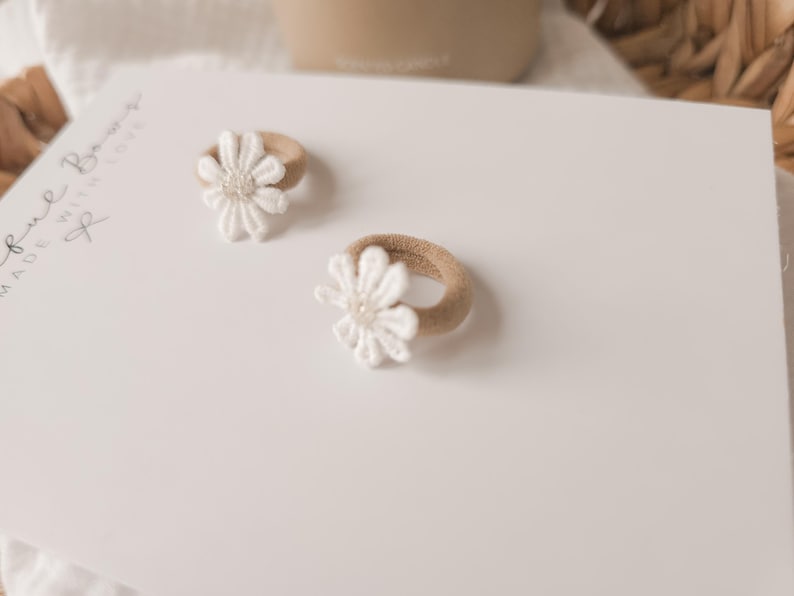 K&ouml;nnte beinhalten: Zwei Baby-Haargummis mit wei&szlig;en G&auml;nsebl&uuml;mchen-Akzenten. Die Haargummis haben ein beiges Gummiband und eine wei&szlig;e Blume mit einem Perlenmittelpunkt. Der Hintergrund ist eine wei&szlig;e Oberfl&auml;che mit den Worten "Soul Bows Made With Love" in Schreibschrift.