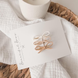 May include: Three small hair bows in white, tan, and light brown are displayed on a white card. The card is printed with the words "Beautiful Bows Handmade with Love". The bows are arranged on a white card, resting on a white cloth.