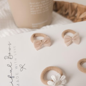 K&ouml;nnte beinhalten: Eine Sammlung von beigefarbenen Baby-Haarschleifen und ein wei&szlig;es Haaraccessoire mit Blume sind auf einer wei&szlig;en Oberfl&auml;che angeordnet. Die Schleifen haben eine weiche Stoffstruktur. Der Text "iful Bows MADE WITH LOVE" ist auf eine wei&szlig;e Karte gedruckt.