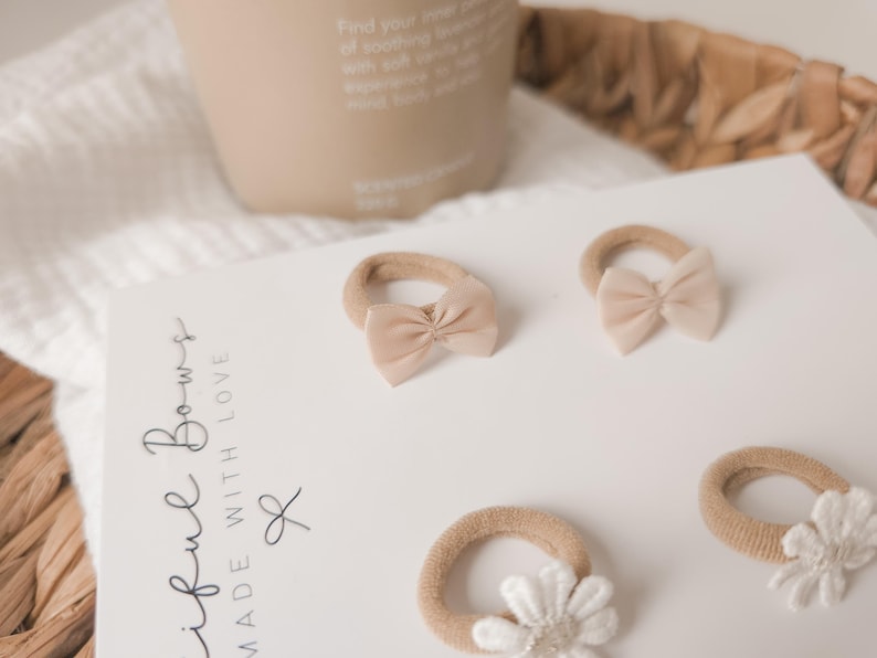 K&ouml;nnte beinhalten: Eine Sammlung von beigefarbenen Baby-Haarschleifen und ein wei&szlig;es Haaraccessoire mit Blume sind auf einer wei&szlig;en Oberfl&auml;che angeordnet. Die Schleifen haben eine weiche Stoffstruktur. Der Text "iful Bows MADE WITH LOVE" ist auf eine wei&szlig;e Karte gedruckt.