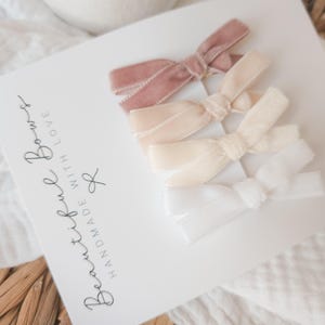 May include: A set of four velvet hair bows in varying shades of pink and white, arranged on a white card. The card features the text "Beautiful Bows, Handmade with Love". The bows are tied in a classic bow shape.