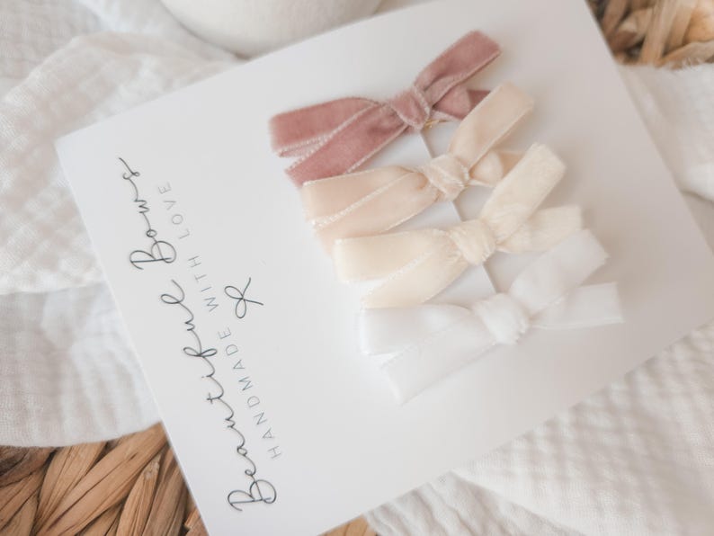 May include: A set of four velvet hair bows in varying shades of pink and white, arranged on a white card. The card features the text "Beautiful Bows, Handmade with Love". The bows are tied in a classic bow shape.