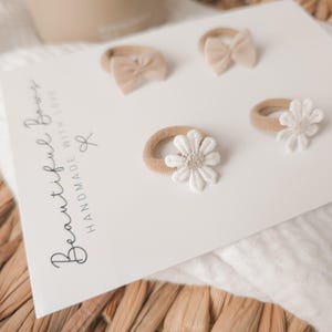 K&ouml;nnte beinhalten: Eine wei&szlig;e Karte zeigt vier Baby-Haaraccessoires. Zwei sind kleine Schleifen und zwei sind g&auml;nsebl&uuml;mchenf&ouml;rmig. Die Accessoires sind in Beige- und Wei&szlig;t&ouml;nen gehalten. Auf der Karte steht "Beautiful Bows Handmade With Love".