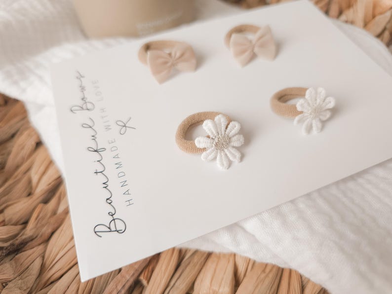 K&ouml;nnte beinhalten: Eine wei&szlig;e Karte zeigt vier Baby-Haaraccessoires. Zwei sind kleine Schleifen und zwei sind g&auml;nsebl&uuml;mchenf&ouml;rmig. Die Accessoires sind in Beige- und Wei&szlig;t&ouml;nen gehalten. Auf der Karte steht "Beautiful Bows Handmade With Love".