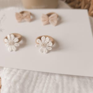 K&ouml;nnte beinhalten: Eine Auswahl an Baby-Haarb&auml;ndern auf einer wei&szlig;en Karte. Zwei Haarb&auml;nder haben kleine Schleifen, und zwei sind mit wei&szlig;en, g&auml;nsebl&uuml;mchenf&ouml;rmigen Verzierungen versehen. Die Haarb&auml;nder sind in neutralen Farbt&ouml;nen gehalten, mit einer weichen, nat&uuml;rlichen &Auml;sthetik.