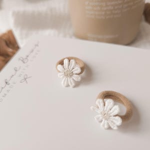 K&ouml;nnte beinhalten: Zwei Baby-Haargummis mit wei&szlig;en G&auml;nsebl&uuml;mchen-Akzenten. Die Haargummis haben ein beiges Gummiband und eine wei&szlig;e, gestickte Blume. Der Text "Beautiful Bows Handmade With Love" ist sichtbar.
