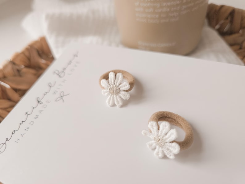 K&ouml;nnte beinhalten: Zwei Baby-Haargummis mit wei&szlig;en G&auml;nsebl&uuml;mchen-Akzenten. Die Haargummis haben ein beiges Gummiband und eine wei&szlig;e, gestickte Blume. Der Text "Beautiful Bows Handmade With Love" ist sichtbar.