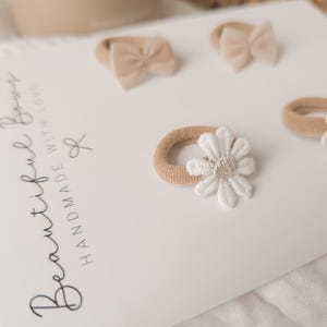 K&ouml;nnte beinhalten: Ein Set aus vier Baby-Haaraccessoires auf einer wei&szlig;en Karte. Zwei Haarspangen haben kleine beige Schleifen, und zwei haben beige B&auml;nder mit wei&szlig;en Blumenakzenten. Auf der Karte steht "Beautiful Bows Handmade With Love".