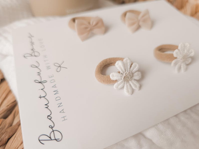 K&ouml;nnte beinhalten: Ein Set aus vier Baby-Haaraccessoires auf einer wei&szlig;en Karte. Zwei Haarspangen haben kleine beige Schleifen, und zwei haben beige B&auml;nder mit wei&szlig;en Blumenakzenten. Auf der Karte steht "Beautiful Bows Handmade With Love".