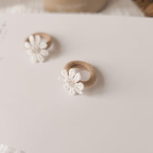 K&ouml;nnte beinhalten: Zwei Baby-Haarb&auml;nder mit wei&szlig;en G&auml;nsebl&uuml;mchen-Akzenten. Die Haarb&auml;nder haben ein beiges Gummiband und eine wei&szlig;e Blume mit strukturiertem Zentrum. Die Haarb&auml;nder liegen auf einer wei&szlig;en Oberfl&auml;che.