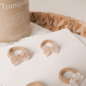K&ouml;nnte beinhalten: Mehrere beige Haargummis mit kleinen, passenden Schleifen sind auf einer wei&szlig;en Oberfl&auml;che angeordnet. Eine Kerze mit dem Wort "Tranquility" ist im Hintergrund zu sehen. Die Haargummis sind in einer neutralen Farbe gehalten.