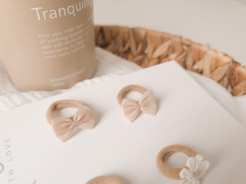 K&ouml;nnte beinhalten: Mehrere beige Haargummis mit kleinen, passenden Schleifen sind auf einer wei&szlig;en Oberfl&auml;che angeordnet. Eine Kerze mit dem Wort "Tranquility" ist im Hintergrund zu sehen. Die Haargummis sind in einer neutralen Farbe gehalten.