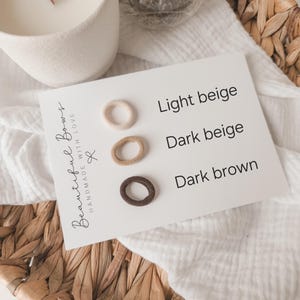 K&ouml;nnte beinhalten: Eine Karte zeigt drei kleine Haargummis in Hellbeige, Dunkelbeige und Dunkelbraun. Auf der Karte steht &bdquo;Hellbeige&ldquo;, &bdquo;Dunkelbeige&ldquo; und &bdquo;Dunkelbraun&ldquo;. Die Karte liegt auf einer wei&szlig;en Oberfl&auml;che mit einem geflochtenen Korb und einer wei&szlig;en Kerze im Hintergrund.