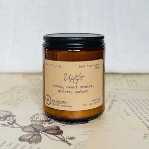 Photo of a glass jar candle with black lid and tan antique style label with script and typewriter fonts. Scented with sweet orange, chocolate, and ginger. Handmade small local business woman owned.