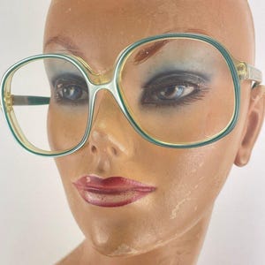 May include: Large, round-framed spectacles in a light green and clear design. The glasses are displayed on a mannequin head. The frames have a retro, vintage aesthetic, reminiscent of the 1970s.