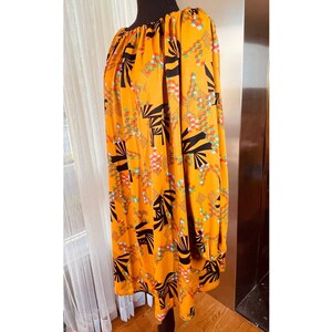 May include: An orange, long-sleeved dress with a black, white, and red geometric pattern. The dress has a round neckline and gathered sleeves. The dress is displayed on a black mannequin.